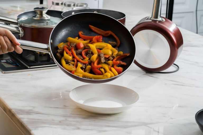 A non-stick pan being used to cook colorful bell peppers and beef, with the food being transferred to a white bowl on a kitchen counter.